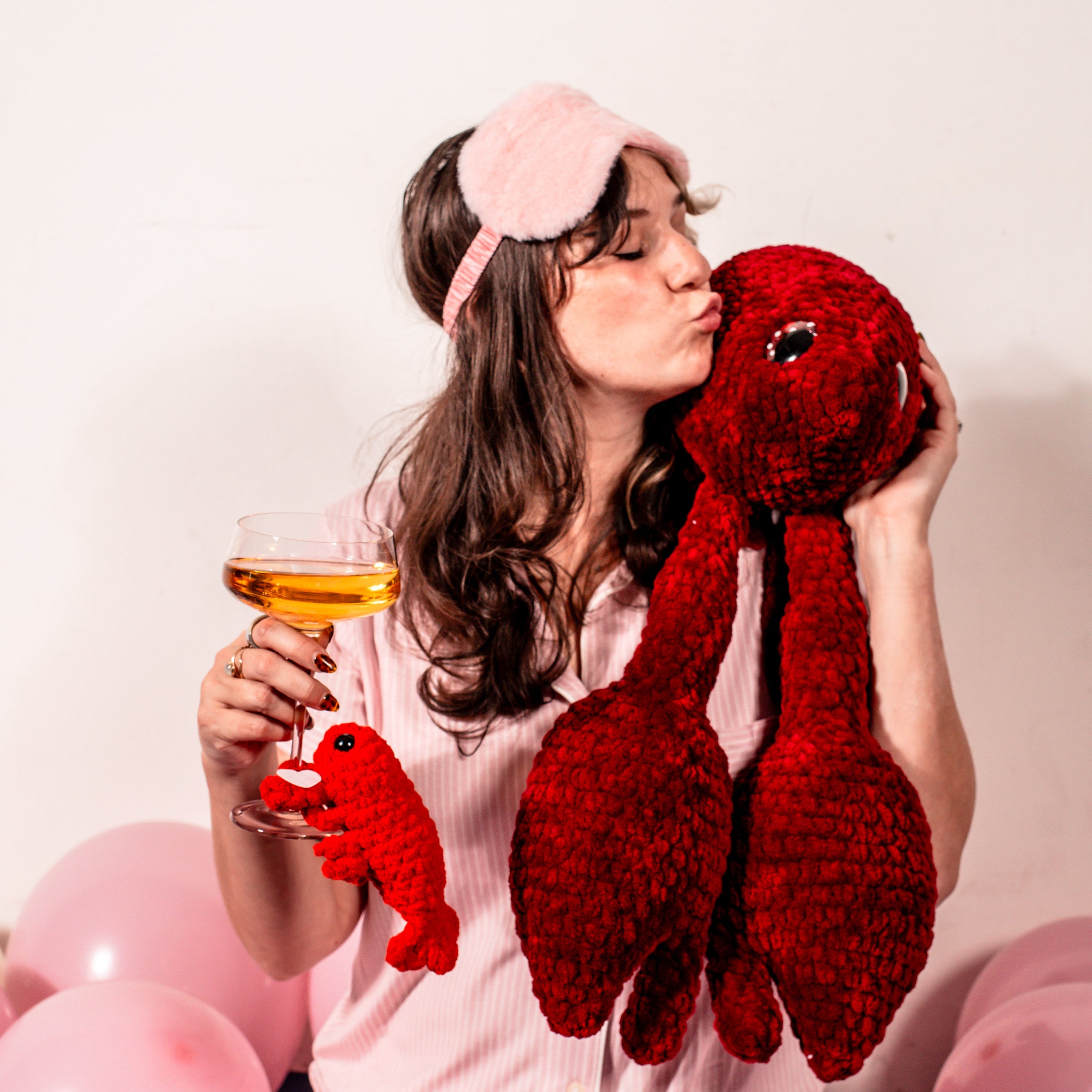 Woman in pajamas holding a glass of wine and a red plush toy, with pink balloons in the background.